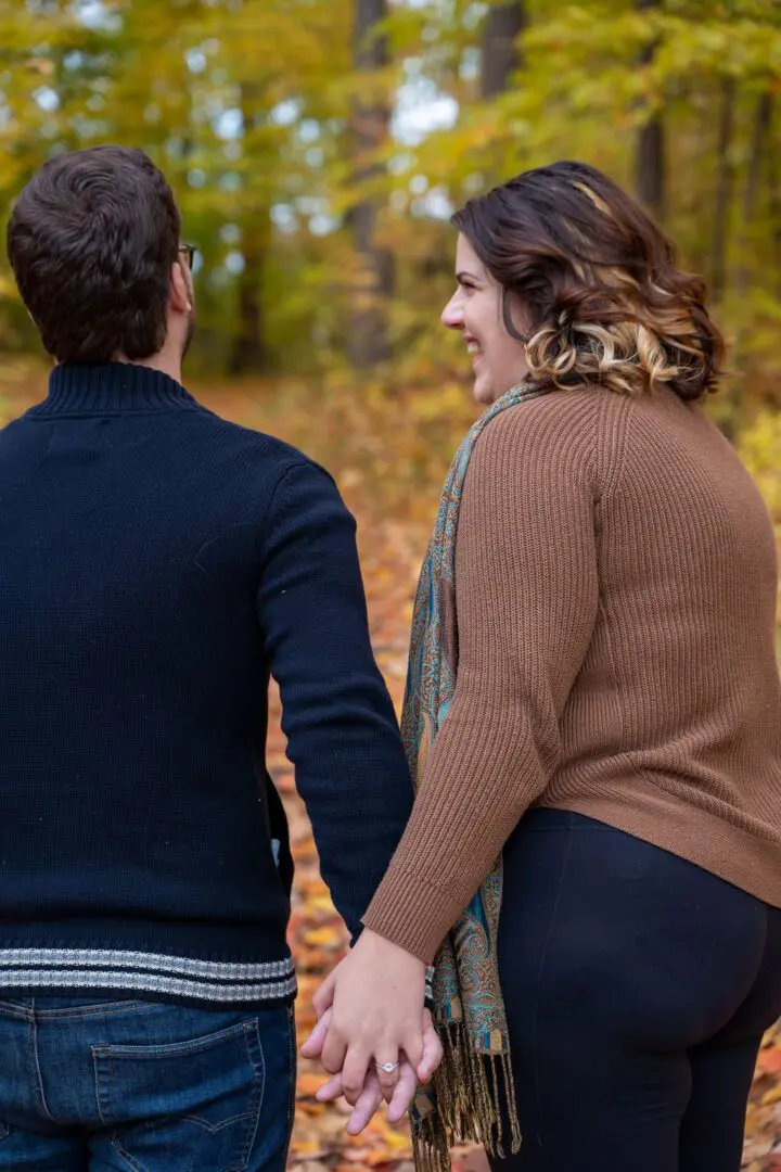 A couple holding hands while walking on a leaf-covered path surrounded by trees with autumn foliage.