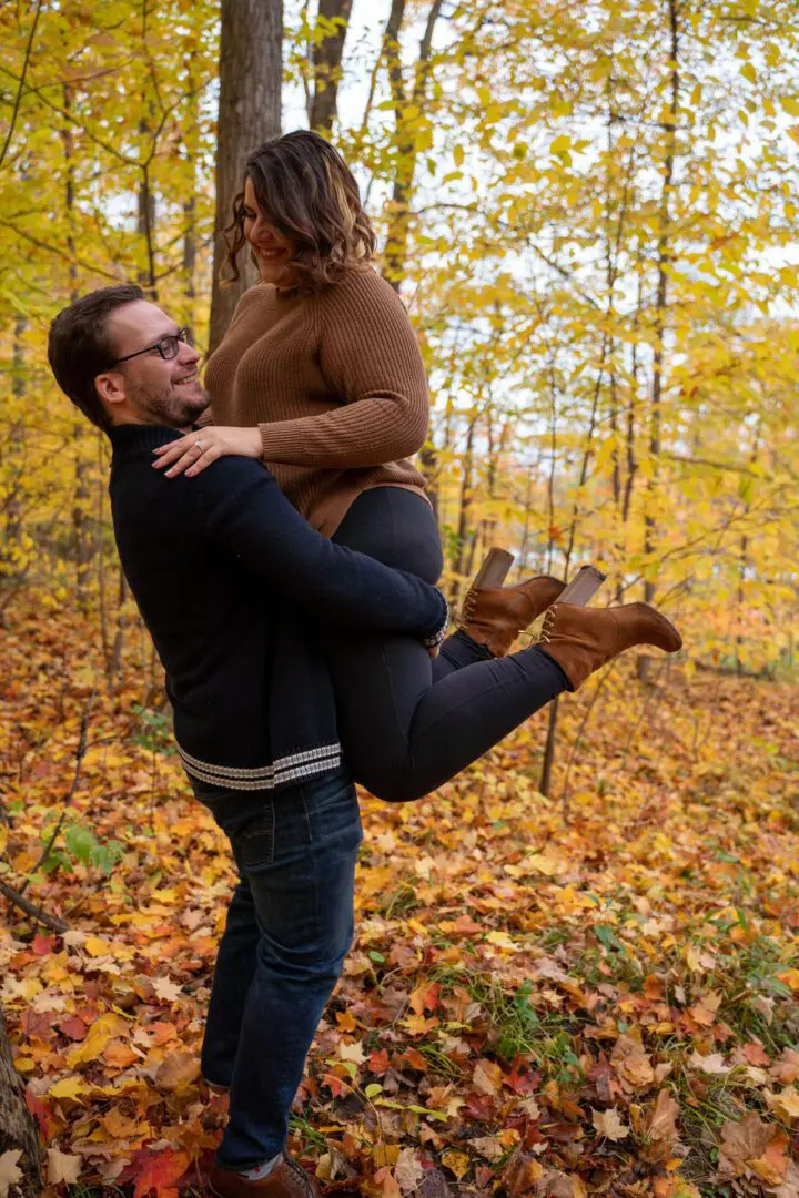 A man lifts a smiling woman in an autumn forest with yellow leaves on trees and ground. Both wear casual fall clothing.