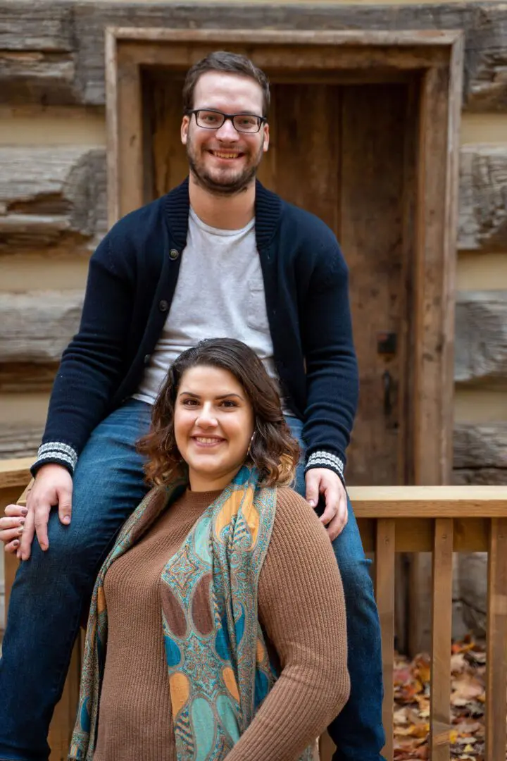 A man sitting on a wooden railing behind a woman in a patterned scarf, both smiling in front of a rustic wooden building.