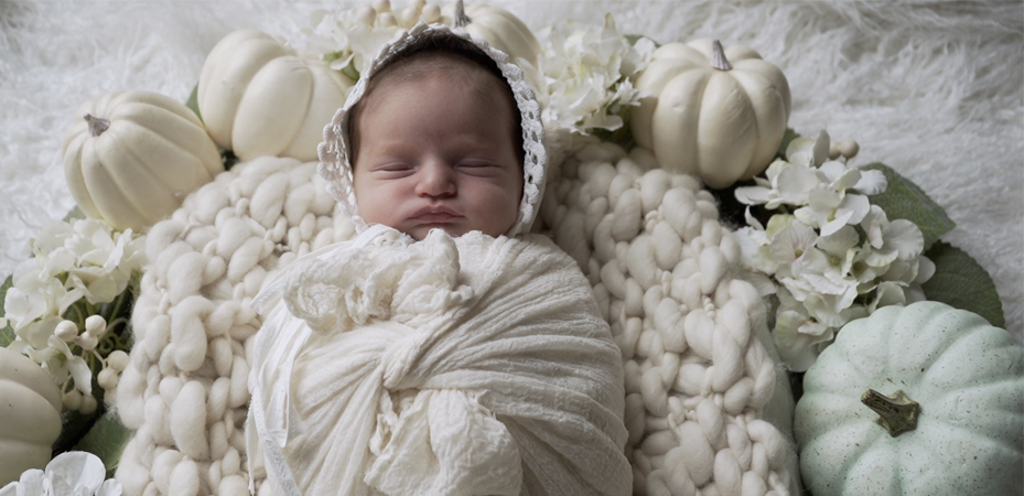 A baby wrapped in white blanket with pumpkins around it.
